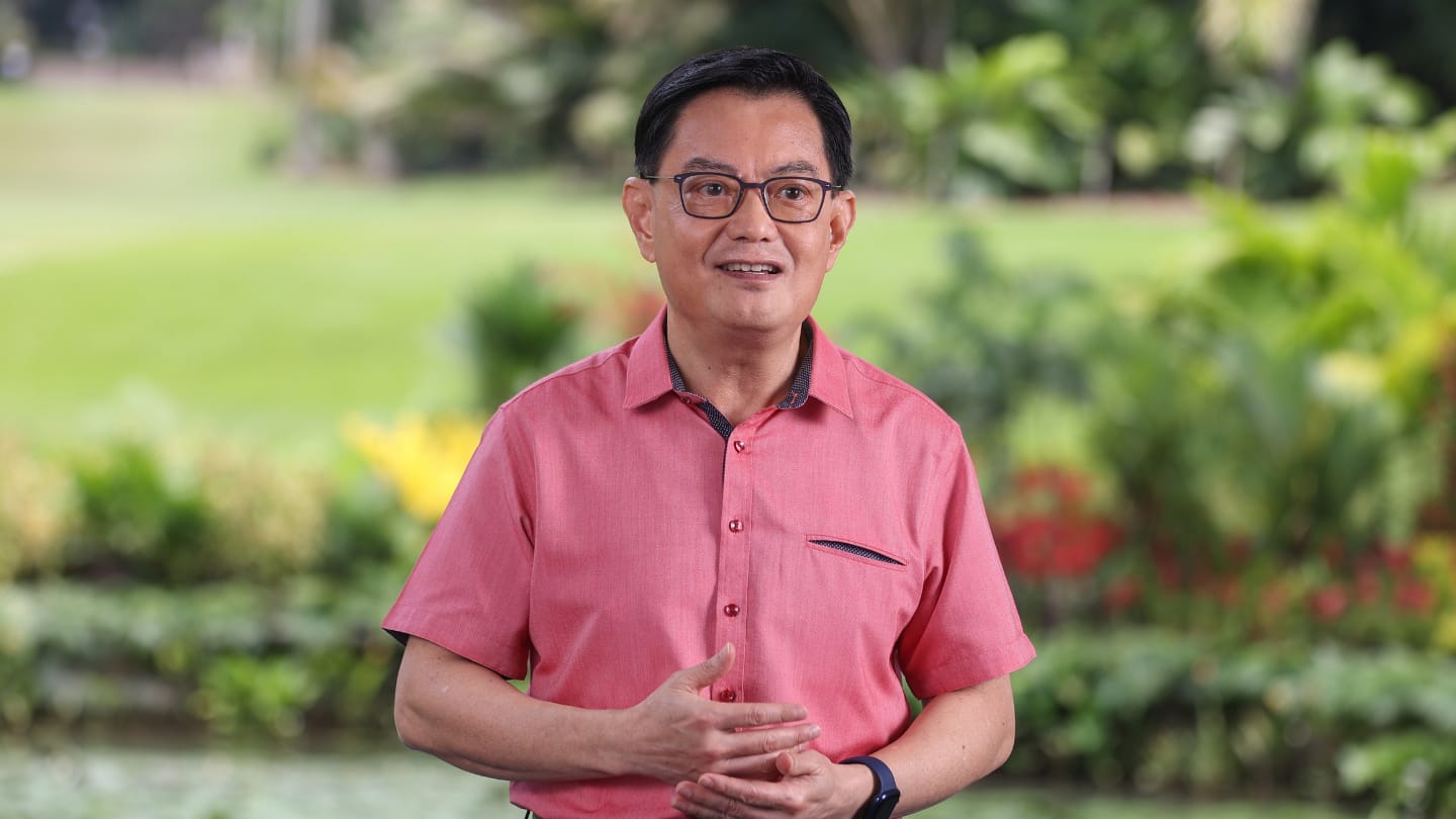 Man in glasses and pink shirt, hands clasped, outdoors with blurred greenery.